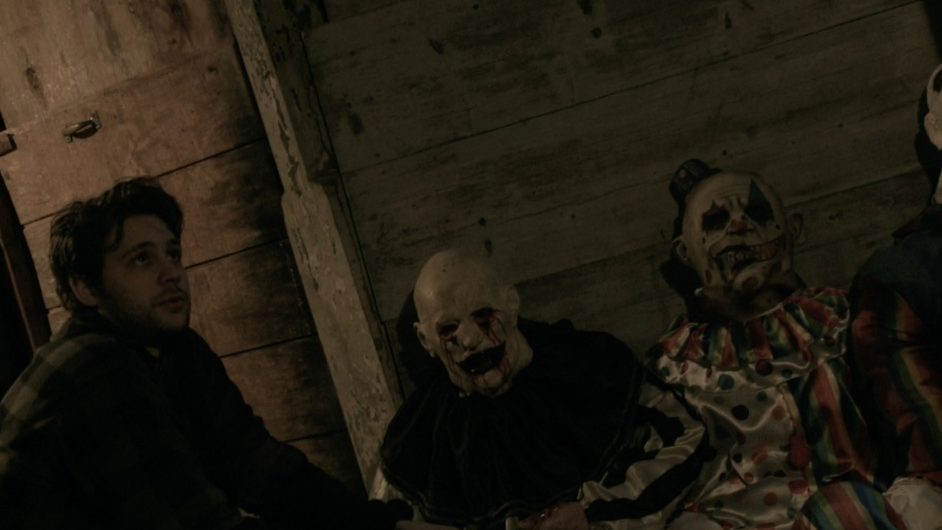 A man crouching next to two clowns during the horror movie, Hell House LLC.