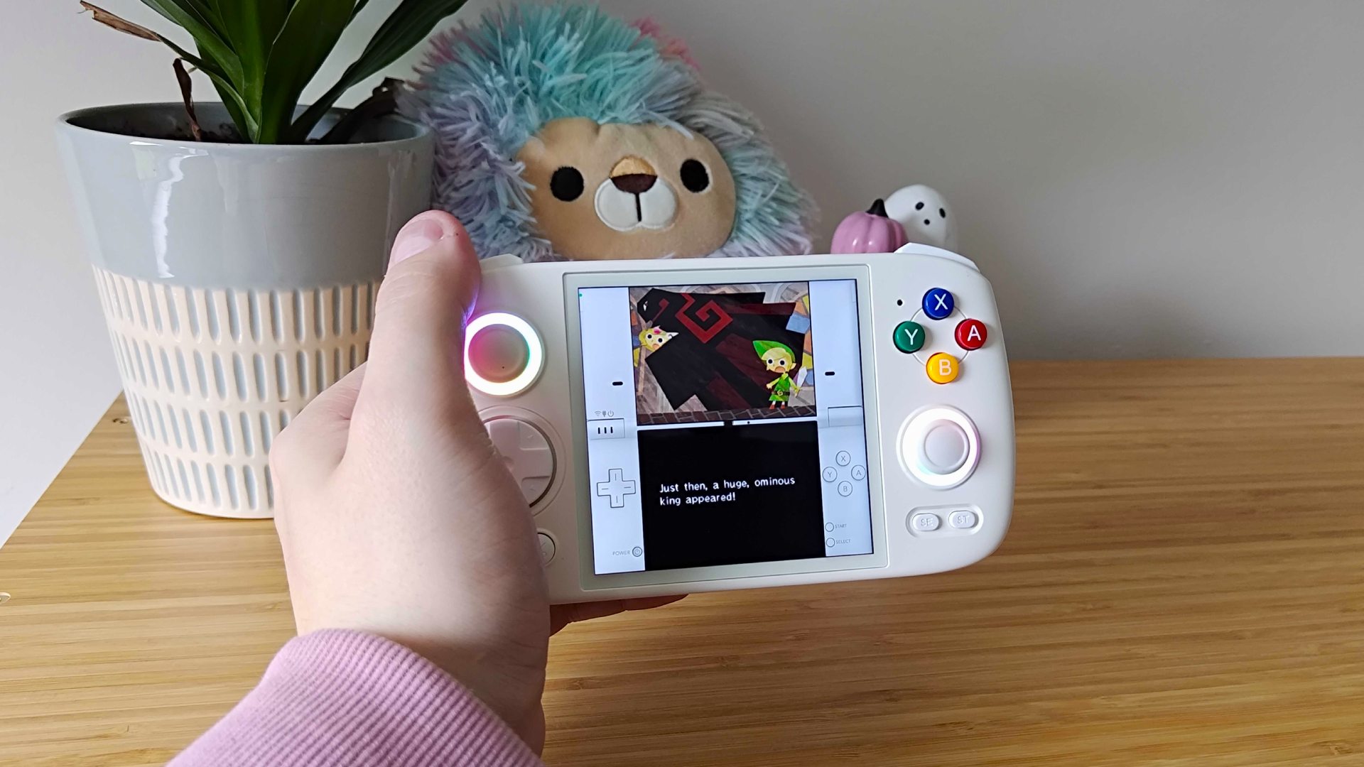 Phil Hayton holding Anbernic RG Cube handheld with plant and Lion Squishmallow in backdrop and Zelda: Phantom Hourglass gameplay on screen