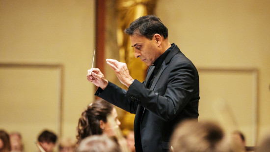 Vijay Upadhyaya stands in profile, his conducting wand raised and his head bowed as he consult the sheet music before him.