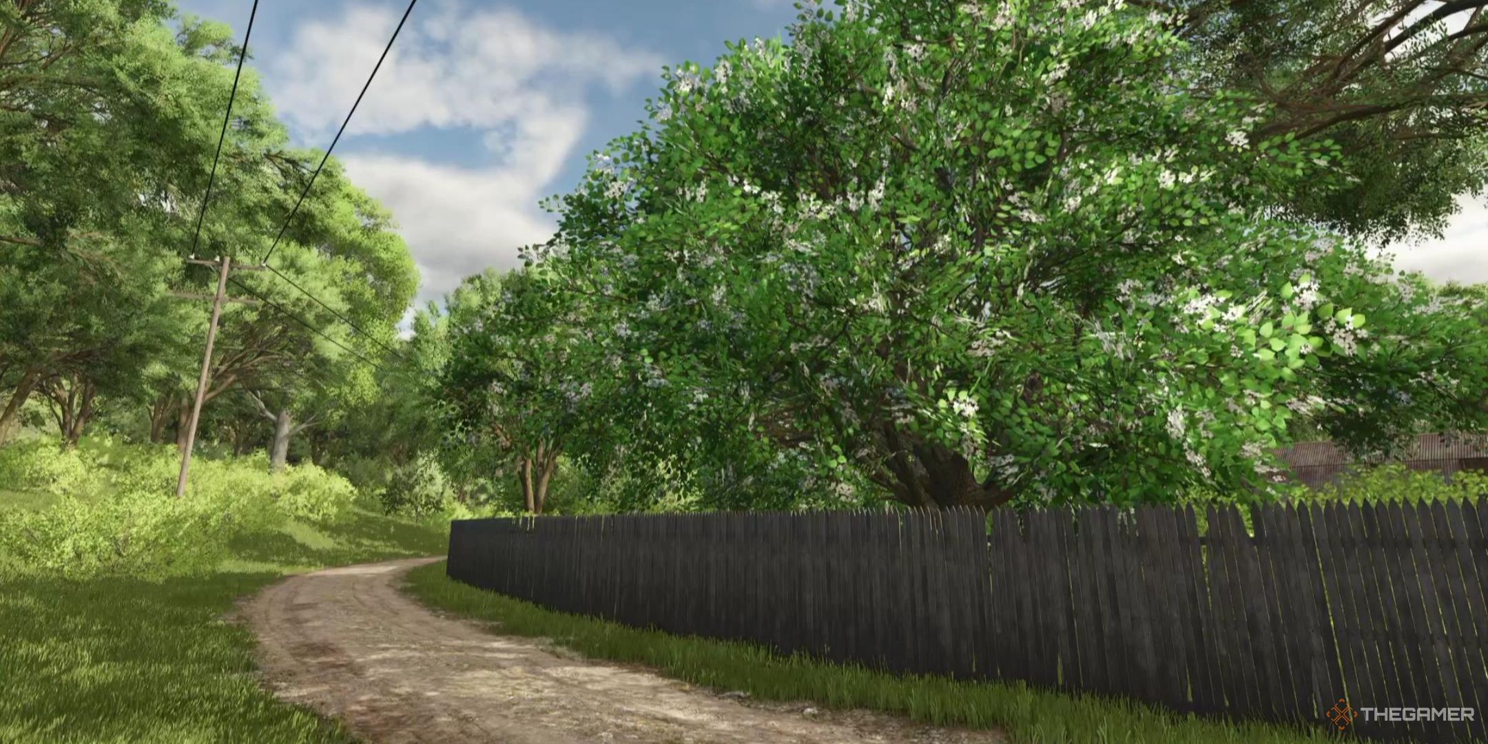 A beautiful, flowering tree sits behind a fence with a dirt road winding off into the distance in Farming Simulator 25.