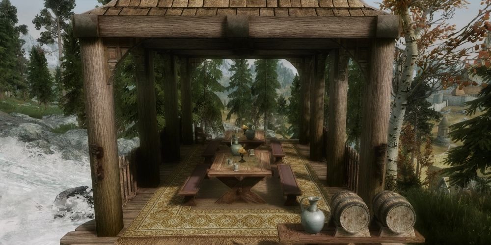 An outdoor room with tables, benches, teapots, teacups, candles, barrels, a jug surrounded by trees and rushing water