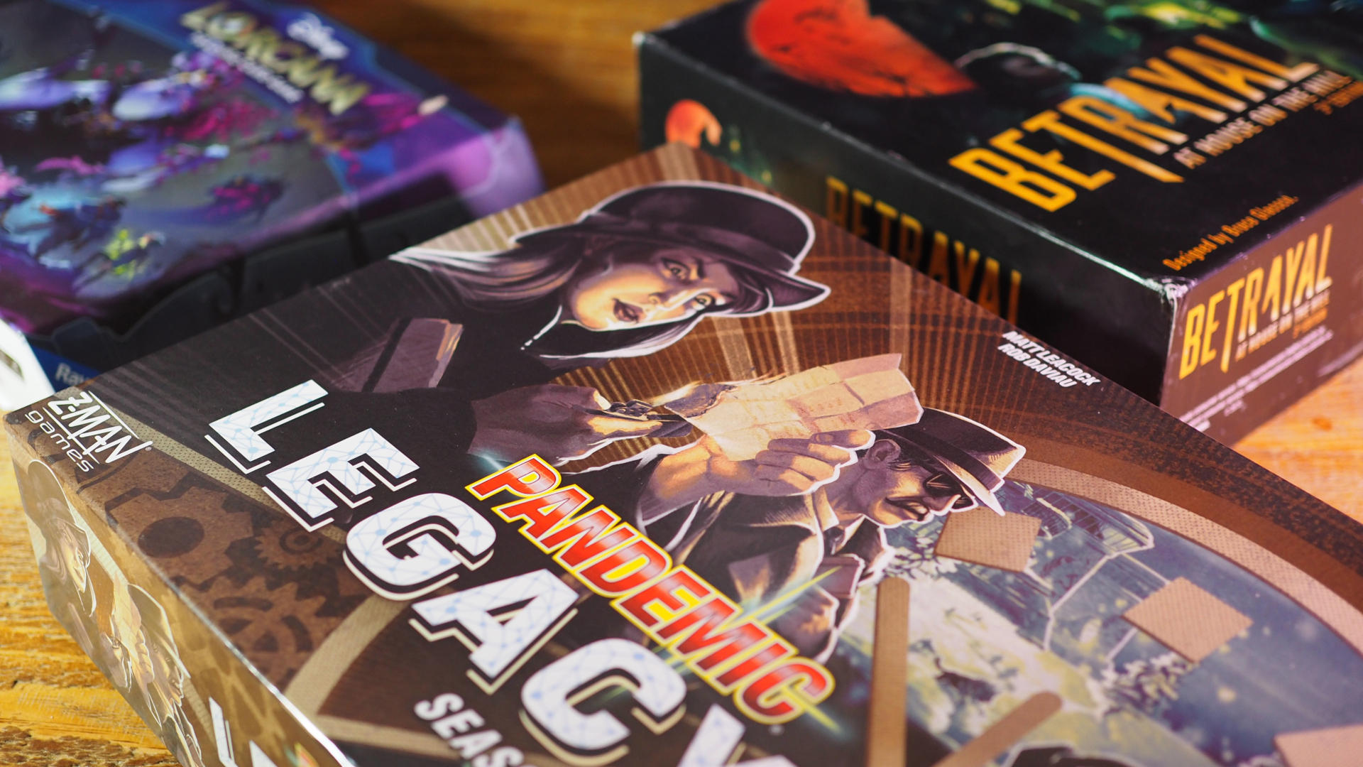 Pandemic Legacy Season 0, Disney Lorcana: Deep Trouble, and Betrayal at House on the Hill 3rd Edition on a wooden table, against a black background