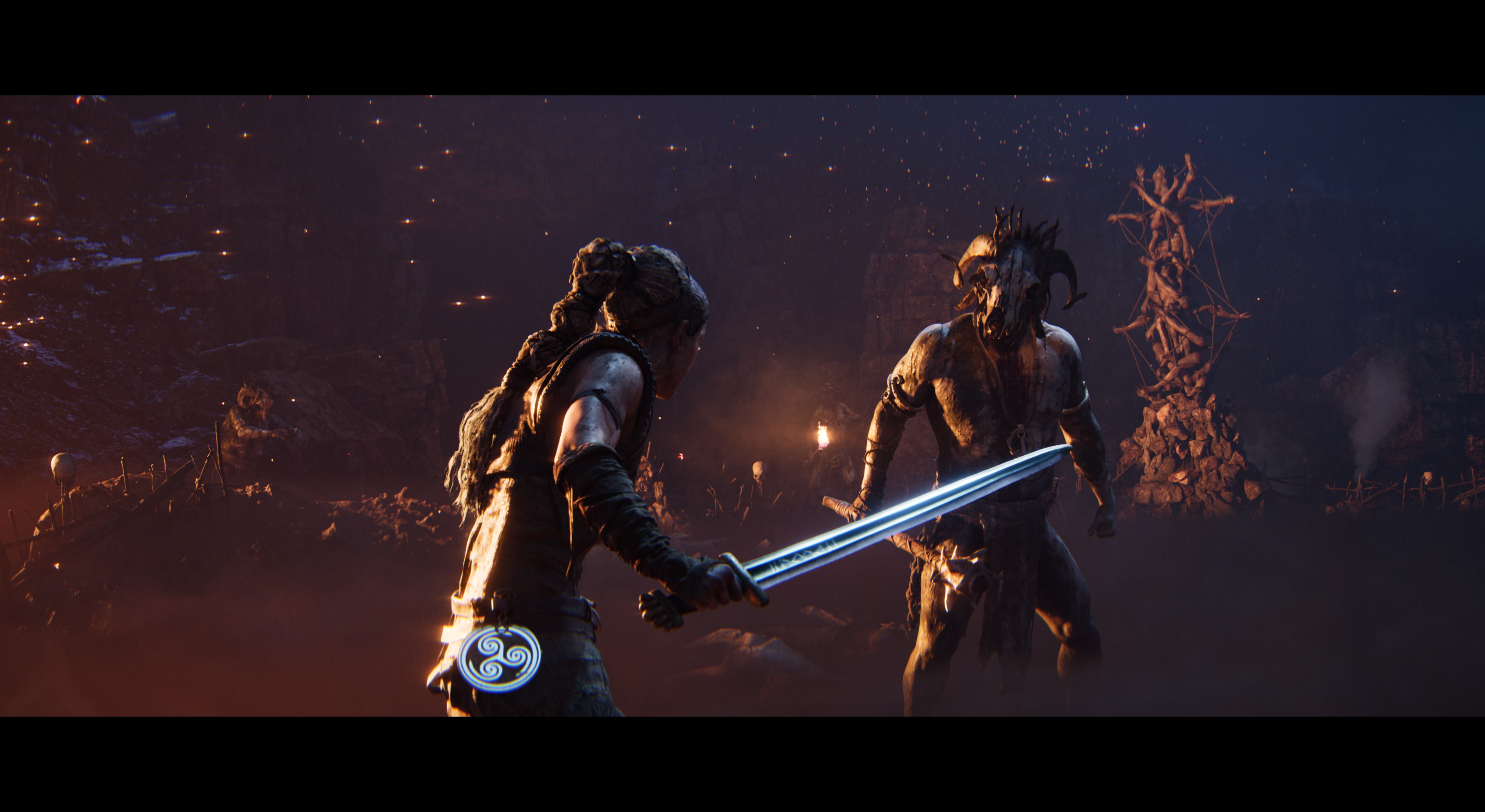 Senua faces down an enemy in a dark scene lit by fire in Senua's Saga Hellblade 2, he is wearing a skull mask, and a tower made up of dismembered limbs is in the background