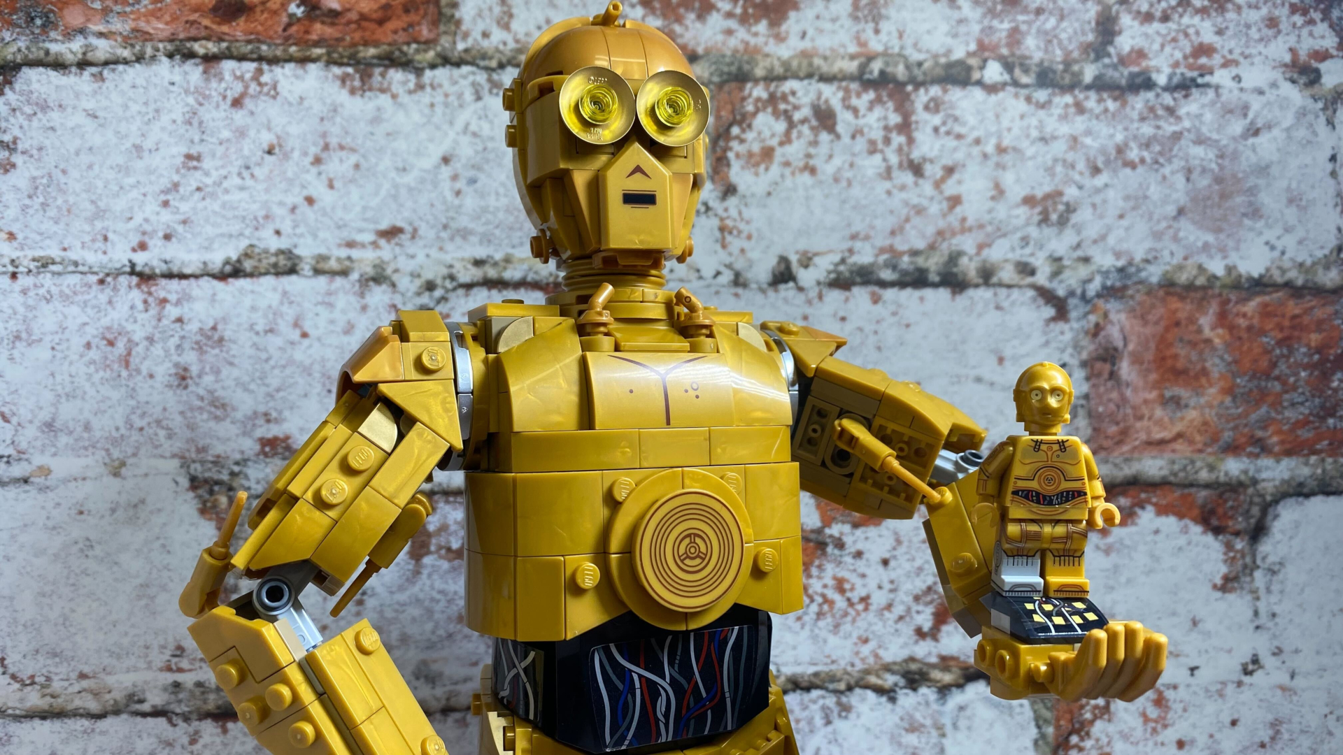 Lego C-3PO holding a smaller Lego C-3PO minifigure, all against a brick wall