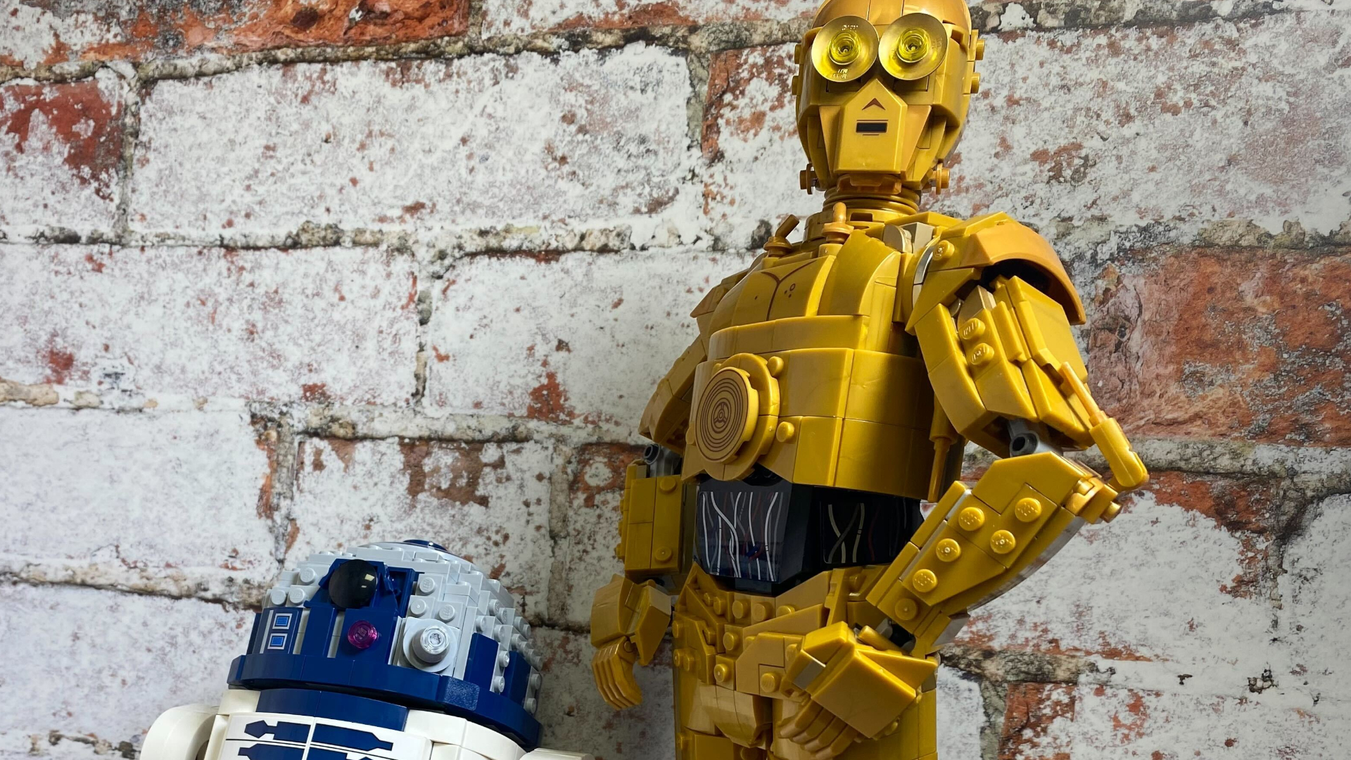 Lego C-3PO and Lego R2-D2 beside each other, against a brick wall