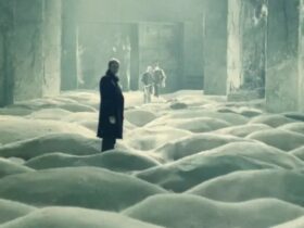 Three men stand in a concrete room where the floor is covered in small dunes of sand.