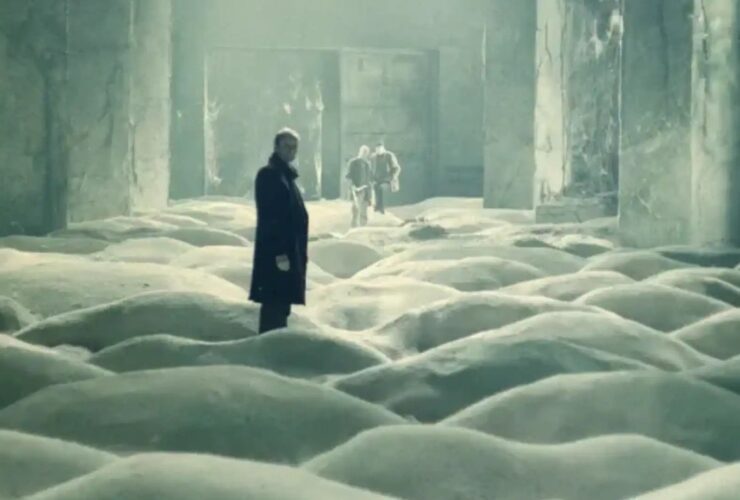 Three men stand in a concrete room where the floor is covered in small dunes of sand.