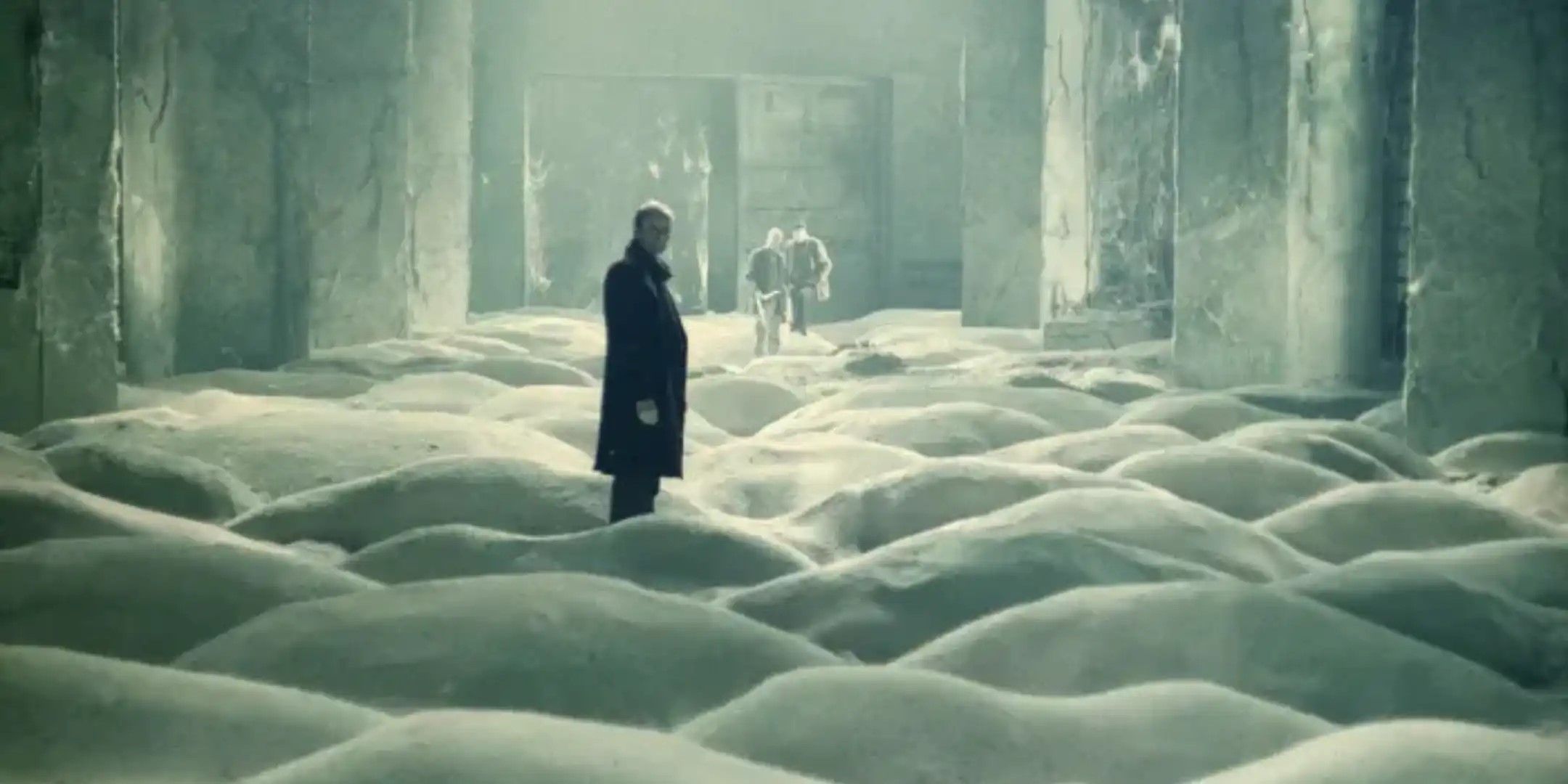 Three men stand in a concrete room where the floor is covered in small dunes of sand.