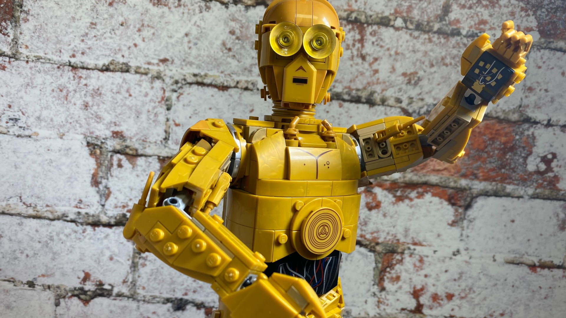 Lego C-3PO holding out an arm, while standing against a brick wall