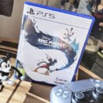 Image of the PS5 copy of Epic Mickey Rebrushed sitting on a coffee table with a Mickey toy and DualSense controller sitting infront of it.