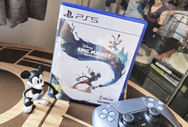 Image of the PS5 copy of Epic Mickey Rebrushed sitting on a coffee table with a Mickey toy and DualSense controller sitting infront of it.