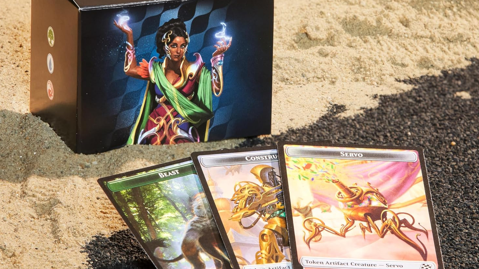 An MTG deck box sits amongst sand, while three token cards lie in front of it on asphalt