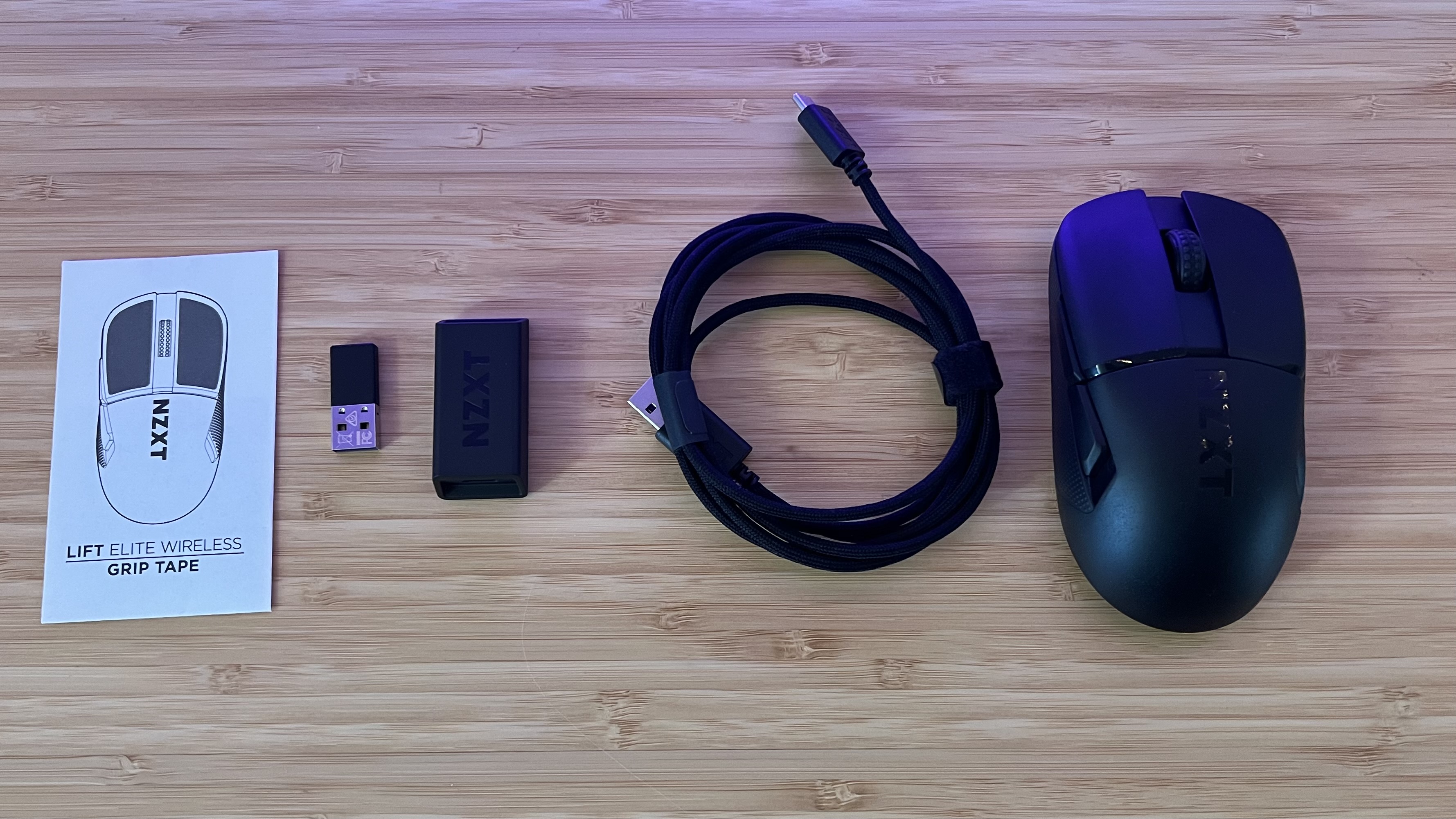 NZXT Lift Elite Wireless gaming mouse with charging cable, USB receivers, and grip tape on a wooden desk