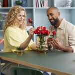 A woman and a man place Lego Bouquet of Roses into a glass vase while sitting at a table