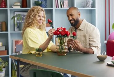 A woman and a man place Lego Bouquet of Roses into a glass vase while sitting at a table