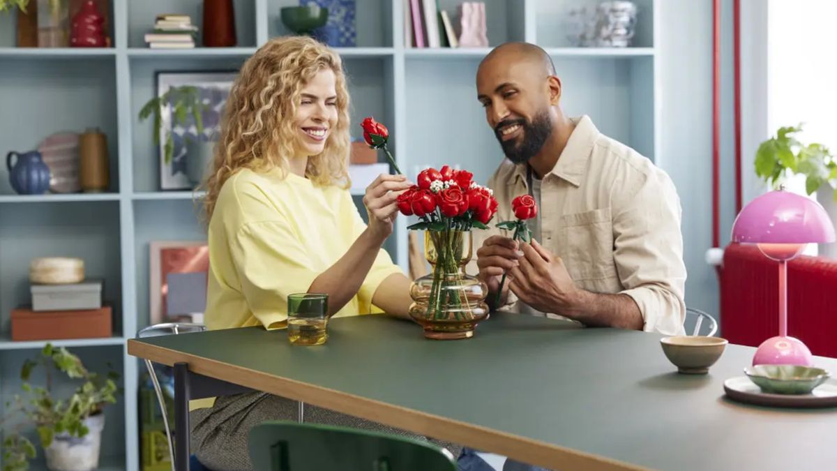 A woman and a man place Lego Bouquet of Roses into a glass vase while sitting at a table