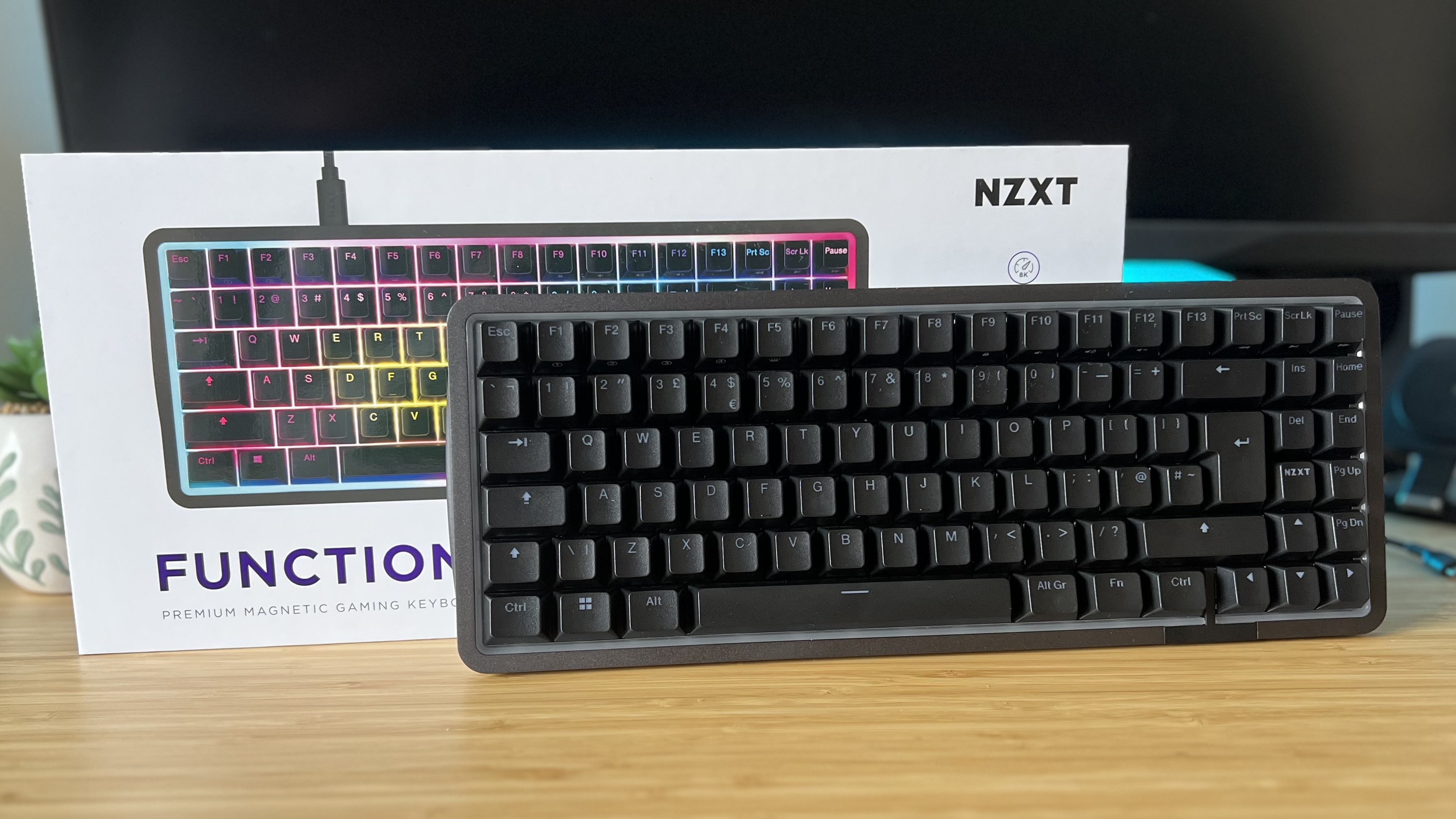 NZXT Function Elite MiniTKL gaming keyboard with RGB off, leaning against packaging on a wooden desk