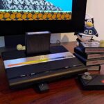 Atari 7800+ console next to stack of games and wireless controller on woodgrain desk