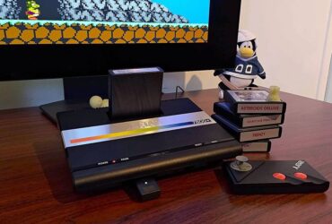 Atari 7800+ console next to stack of games and wireless controller on woodgrain desk