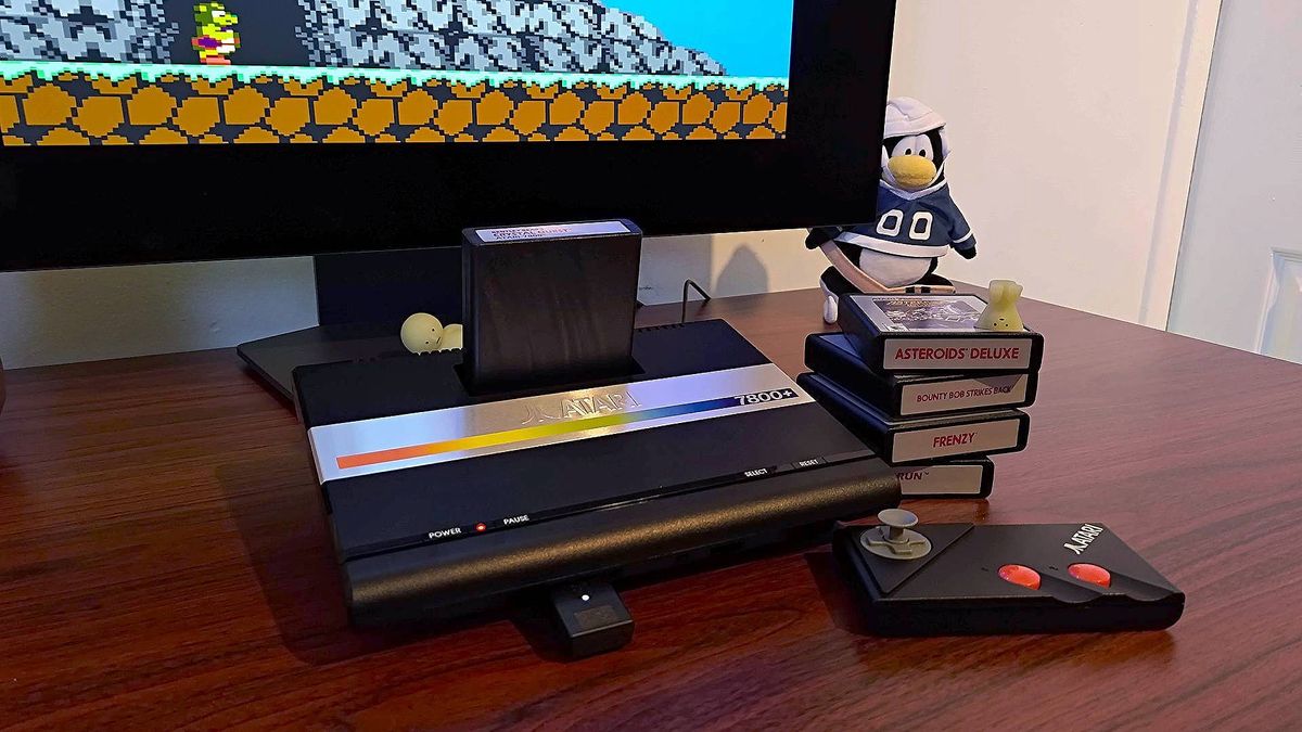 Atari 7800+ console next to stack of games and wireless controller on woodgrain desk