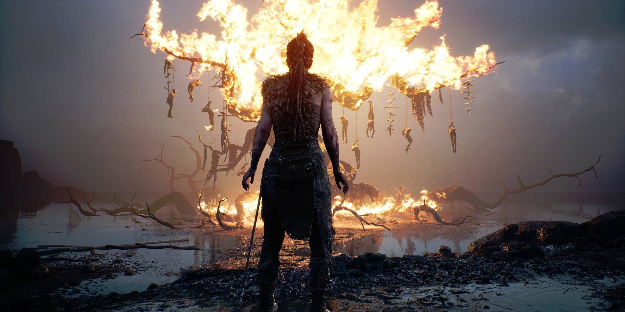 Senua stands in front of a burning tree on a dying landscape in Hellblade Senua's Sacrifice.