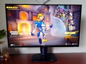 Alienware AW2725Q gaming monitor sitting on woodgrain desk next to plant with Marvel Rivals on screen and Invisible Woman selected on character screen.