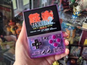 Photo by writer Rosalie Newcombe of their hand holding up the Atomic Purple Miyoo Mini Plus retro gaming handheld infront of a cabinet full of anime toys and figures.