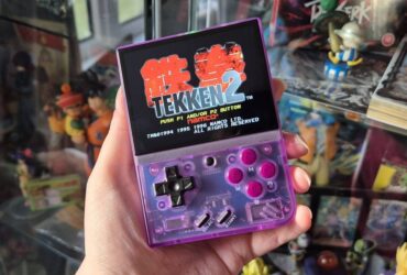 Photo by writer Rosalie Newcombe of their hand holding up the Atomic Purple Miyoo Mini Plus retro gaming handheld infront of a cabinet full of anime toys and figures.