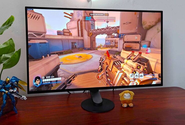 Sony Inzone M10S gaming monitor sitting on woodgrain desk next to plant and Pharah figure with Overwatch 2 gameplay in screen.