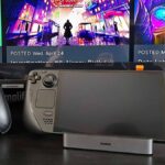 Baseus 6-in-1 Steam Deck dock with OLED model sitting in cradle next to Asus controller and Samsung TV with game menu in backdrop.