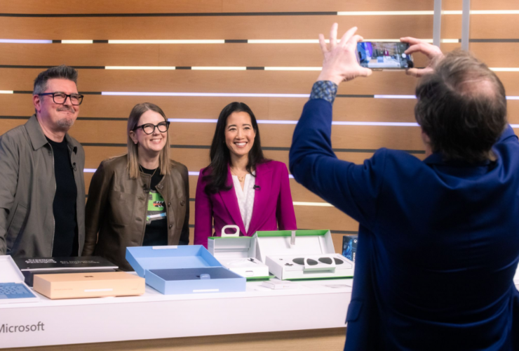 Four people are in the image. Three of them are standing behind a table, and one person is taking a photo of them. The person taking the photo is wearing a blue suit and holding a smartphone. The three people behind the table are smiling. The man on the left is wearing glasses and a brown jacket over a black shirt. The woman in the middle is wearing glasses and a brown jacket over a black shirt with a green design. The woman on the right is wearing a magenta blazer over a white top. The table has several boxes on it, and the word "Microsoft" is visible on the side. The background consists of horizontal wooden panels.