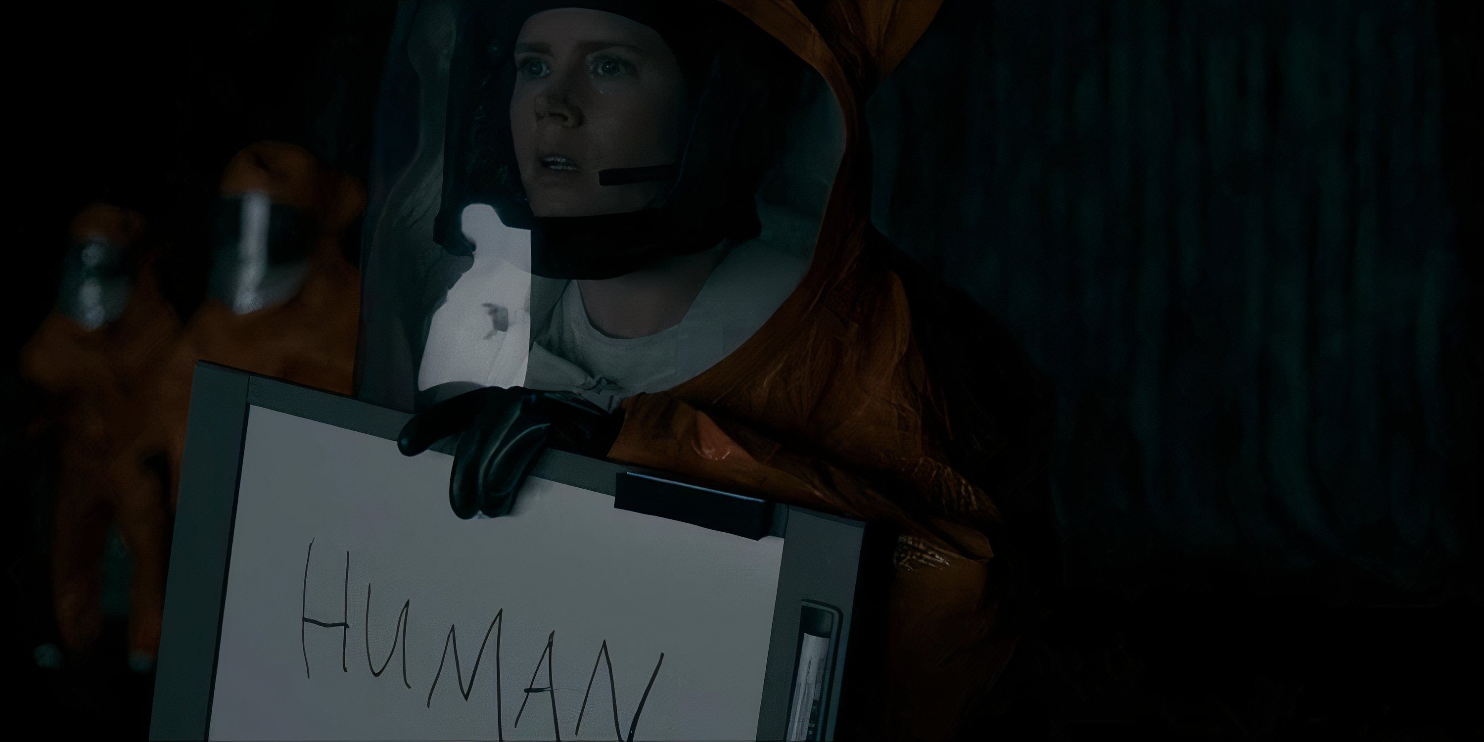 Amy Adams in an orange hazmat suit holding up a sign that communicates to the aliens that she's human in Arrival.