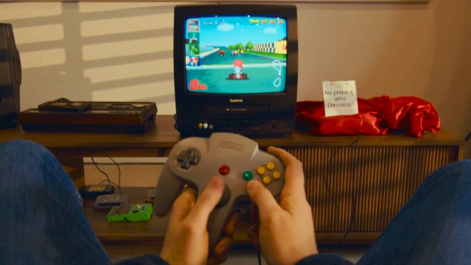 Player holding N64 controller in front of CRT with Mario Kart 64 on screen and console covered in red cloth next to sign that reads &quot;no peeking until Christmas!&quot;