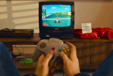 Player holding N64 controller in front of CRT with Mario Kart 64 on screen and console covered in red cloth next to sign that reads &quot;no peeking until Christmas!&quot;